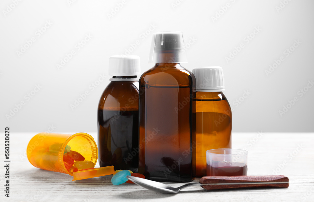 Bottles of syrup, measuring cup, lozenges and spoon on white wooden table against light grey background. Cold medicine Bottles of syrup, measuring cup, lozenges and spoon on white wooden table against light grey background. Cold medicine
