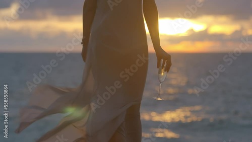 Woman walks, dressed in light summer silk dress, against sunset on the ocean beach, holds a glass of champagne