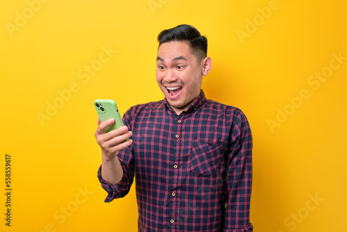 Surprised young Asian man looking at smartphone screen and emotionally reacting to online news isolated over yellow background
