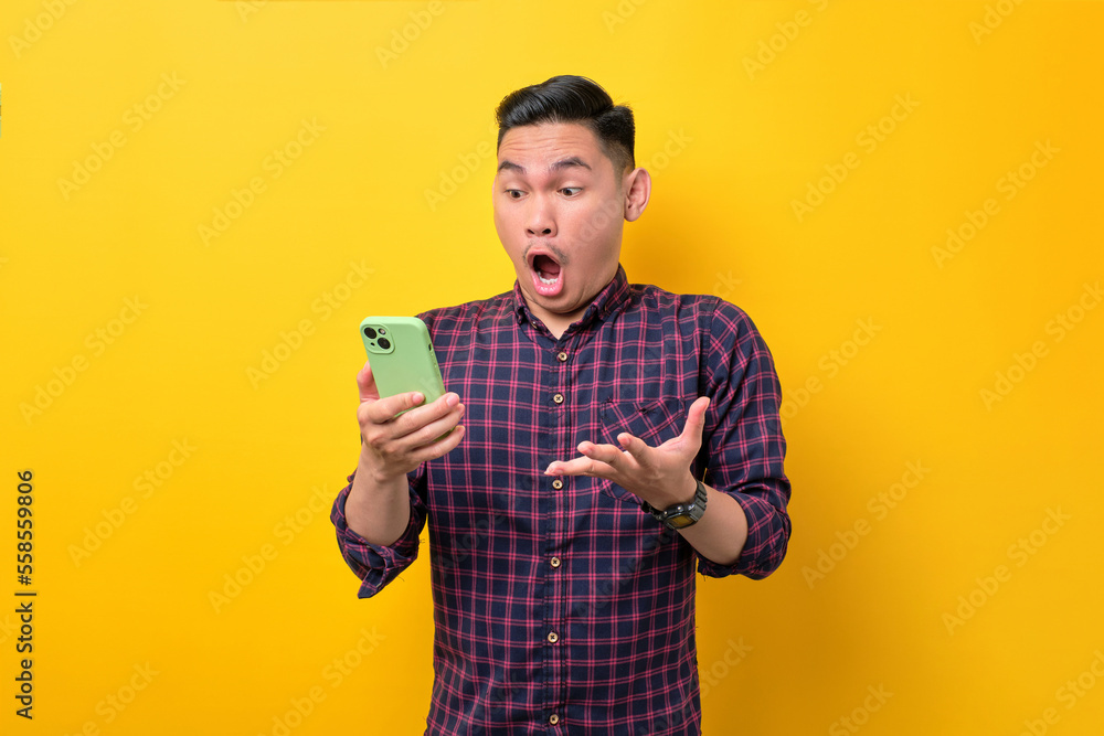 Surprised young Asian man looking at smartphone screen and emotionally reacting to online news isolated over yellow background Surprised young Asian man looking at smartphone screen and emotionally reacting to online news isolated over yellow background