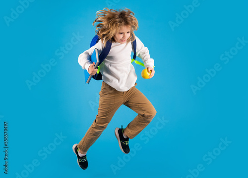 Kid jump and enjoy school. Full body of energetic primary school kid boy wearing casual clothes holding backpackand copybook and jumping against blue background.