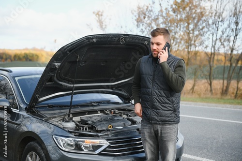 Man repairing a broken car by the road. Man having trouble with his broken car on the highway roadside. Man looking under the car hood. Car breaks down on the autobahn. Roadside assistance concept.