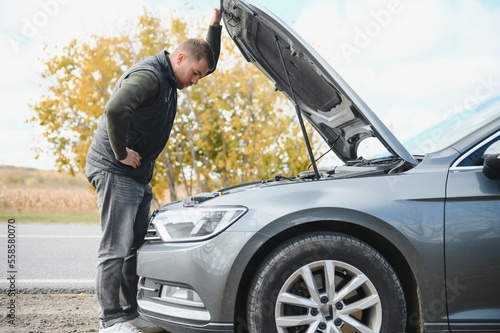 Man repairing a broken car by the road. Man having trouble with his broken car on the highway roadside. Man looking under the car hood. Car breaks down on the autobahn. Roadside assistance concept. © Serhii