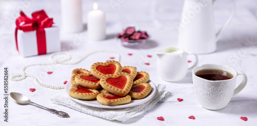 Homemade cookies in the form of hearts with red jam on Valentine's Day. On a marble table with a gift, tea and white candles.