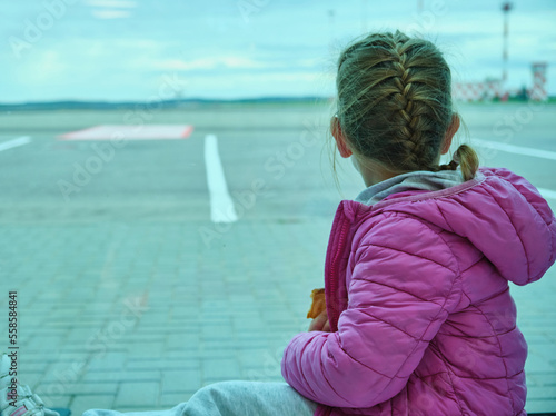 Little girl looking waiting through window to airplane in airport pink jacket