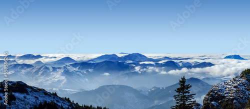 An epic ocean of clouds and fog in the winter mountains landscape, aerial view. Huge white clouds come in waves over the foggy valleys, mountain peaks rise over like islands. Dramatic overcast sky