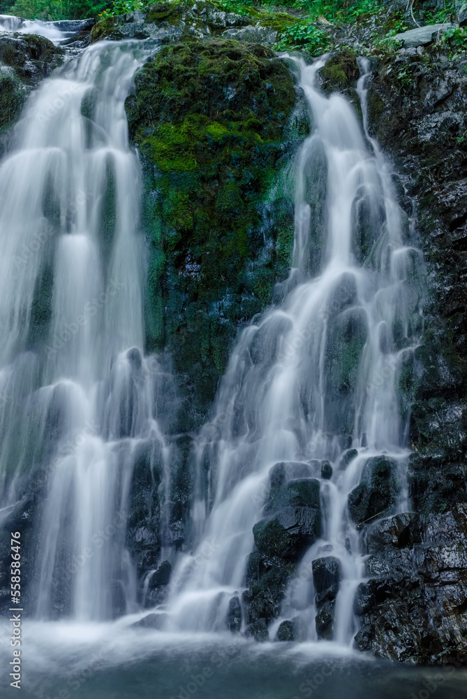 Beautiful mountain rainforest waterfall with fast flowing water and rocks, long exposure. Natural seasonal travel outdoor background with sun shining. Stream waterfall on rocks in the forest