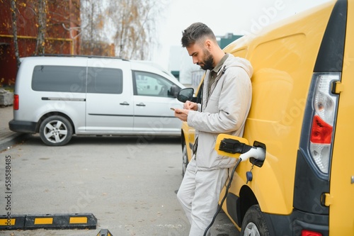 bearded caucasian man standing near an electric car that is charging and making time adjustments on a smartphone. © Serhii