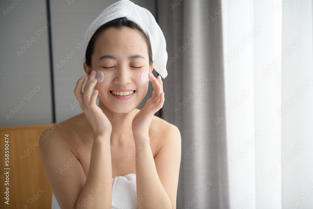 Happy young Asian woman applying face lotions while wearing a towel and touching her face. Daily makeup and skincare Happy young Asian woman applying face lotions while wearing a towel and touching her face. Daily makeup and skincare