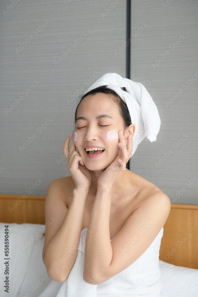 Happy young Asian woman applying face lotions while wearing a towel and touching her face. Daily makeup and skincare Happy young Asian woman applying face lotions while wearing a towel and touching her face. Daily makeup and skincare