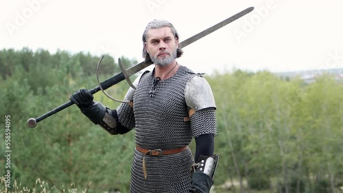 Portrait of a medieval warrior with gray hair and a scar on his face in chain mail with shoulder pads and a two-handed sword in his hands posing against the backdrop of the forest. Witcher Middle Ages