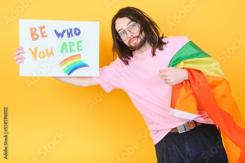 Young queer bisexual smiling gay man with make up in beige tank shirt hold card sign with be who you are title text on rainbow flag background studio portrait.