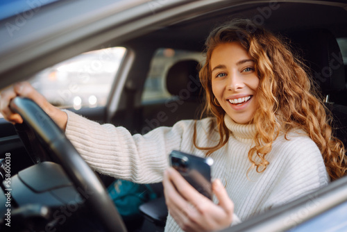 Beautiful young woman is sitting inside a car on the drivers seat with phone. Business, taxi, technologie, internet, online concert.