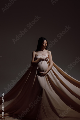 Full length of a Caucasian woman who is pregnant, wearing a long beige cloth on a dark background, space for text photo