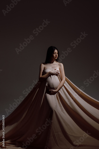 Full length of a Caucasian woman who is pregnant, wearing a long beige cloth on a dark background, space for text
