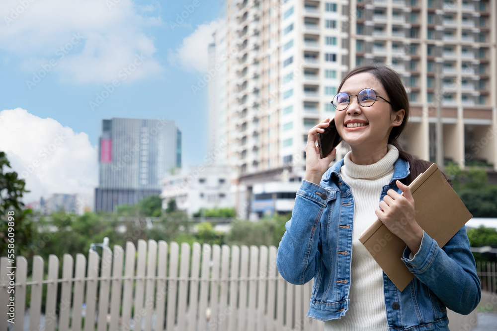 Beautiful young woman in a city. Young Asian woman wearing glasses using smartphone with smiling confident holding book at city street on blue sky. Successful concept
