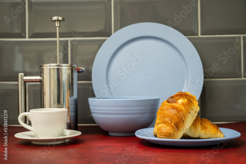 expresso coffee cup and saucer stainless steel cafetiere   with fresh coissant blue dinner set  on red linoleum work top with grey tiled splash back photo