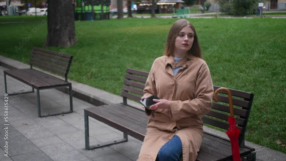 Young beautiful woman sitting on bench and using mobile phone outdoors, then she opening umbrella and walking away when it starts raining Young beautiful woman sitting on bench and using mobile phone outdoors, then she opening umbrella and walking away when it starts raining