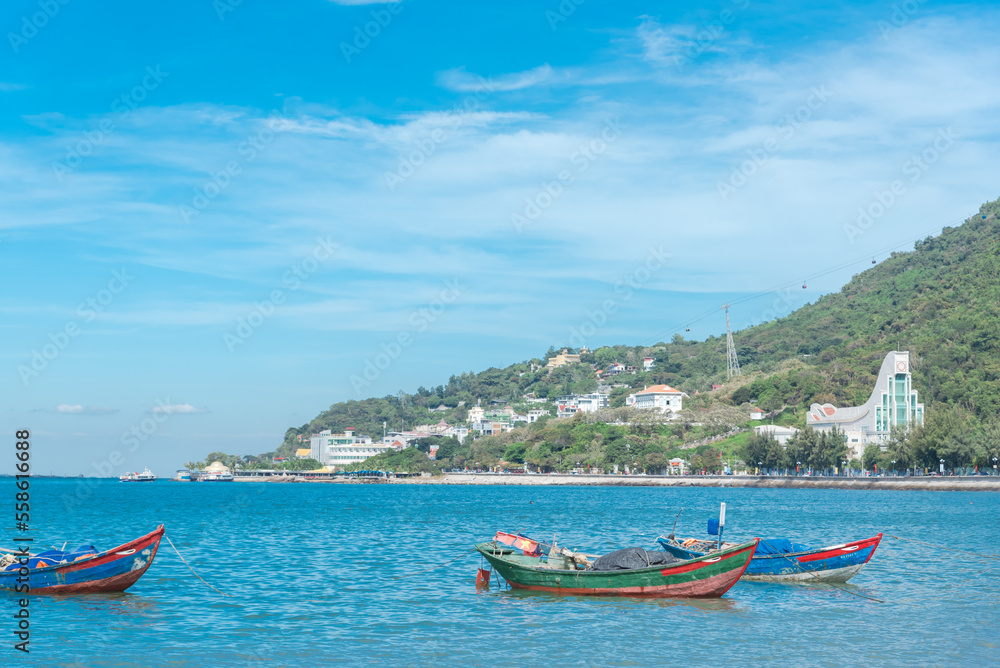 Wooden fishing boats with Vietnamese flags anchored near shoreline at Bai Truoc beach with mountain cable cars and hotel buildings in background, Vung Tau, Vietnam