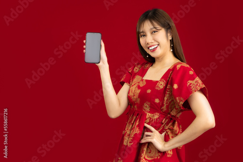 Young beautiful asian woman long hair style in red chinese dress holding blank black screen mobile looking camera red backgroud.