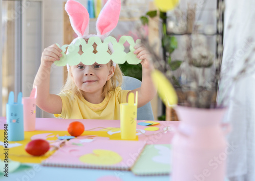 Happy Easter. A beautiful three-year-old girl dressed in bunny ears sitting at a table and holding homemade paper Easter bunnies Garland