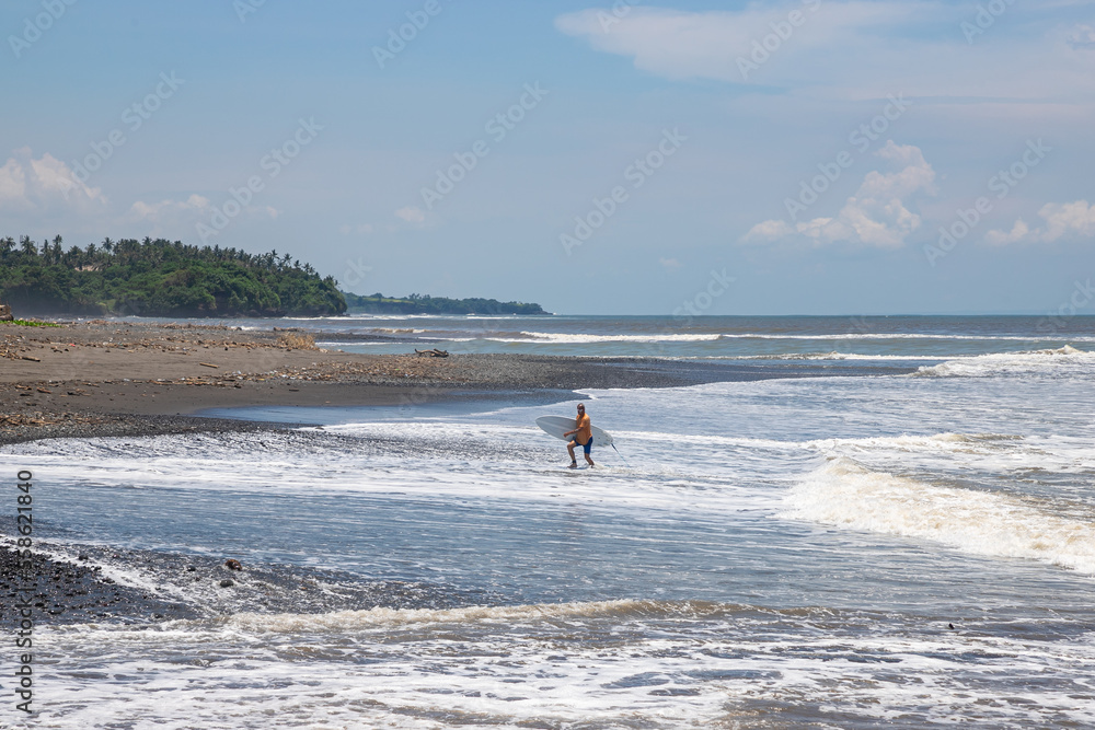 BALI, INDONESIA - NOVEMBER 10, 2022: Male surfer with surfboard spending sunny day on beach near waving ocean in Bali