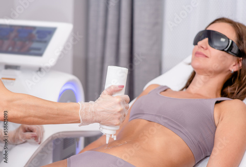 Beautician applying gel on female tummy before epilation. Cosmetologist preparing woman skin for laser hair removal treatment in cosmetology clinic