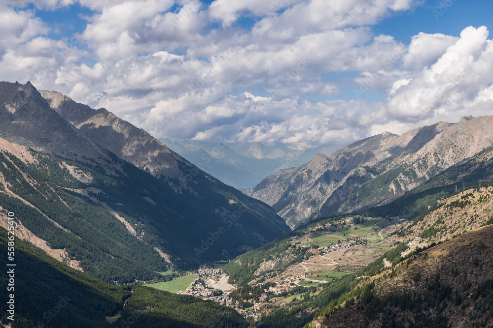 Panoramic shot of alpine gorge with green vegetation on slopes, hikers winding route passes through Cogne village in lowlands, cumulus clouds above, Aosta Valley, Italy