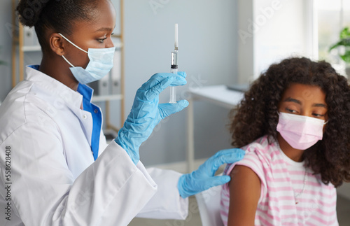 Doctor african american woman is giving a vaccine injection to teen girl in shoulder in clinic. Healthcare, vaccination from flu, covid-19, immunization concept. They wearing protective medical masks.