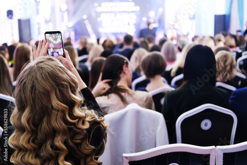 The girl photographs the event on the phone. Guests in evening attire sit in a spotlighted lobby and look at the stage.