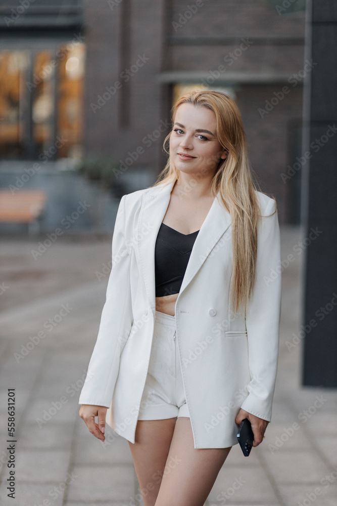 Blond stylish woman at oversized white jacket and phone on hand posing at the railing and steps on the city background