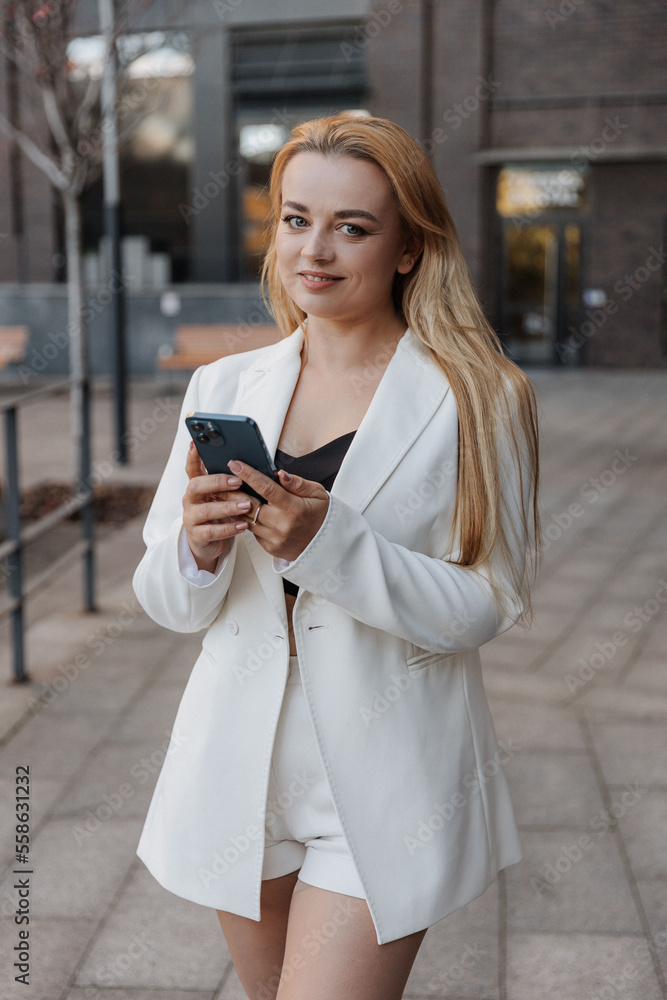 Blond stylish woman at oversized white jacket using phone on the modern city background Blond stylish woman at oversized white jacket using phone on the modern city background