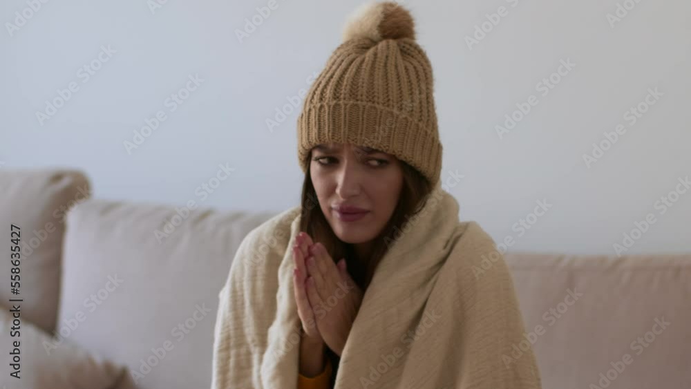 Young cold woman wrapped in blanket with knitted hat on her head freezing at home, warming up her hands and shivering Young cold woman wrapped in blanket with knitted hat on her head freezing at home, warming up her hands and shivering