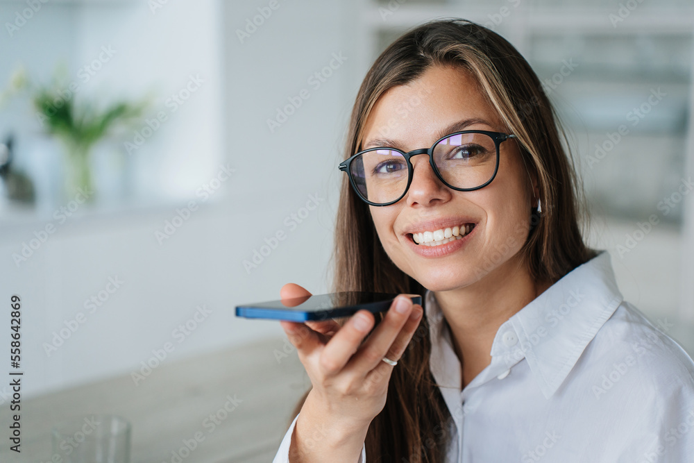 Cheerful hispanic girl in white shirt holding phone recording audio message looks at camera broad smiling against blurry interior. Successful young entrepreneur talks by cell phone using microphone. Cheerful hispanic girl in white shirt holding phone recording audio message looks at camera broad smiling against blurry interior. Successful young entrepreneur talks by cell phone using microphone.