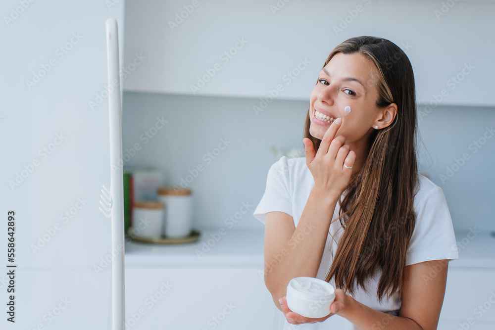 Pretty Italian young woman with long loose hair standing at living room puts skin cream on face broad smiles satisfied by skin care products and healthy lifestyle. Cosmetics. Satisfied female at home. Pretty Italian young woman with long loose hair standing at living room puts skin cream on face broad smiles satisfied by skin care products and healthy lifestyle. Cosmetics. Satisfied female at home.