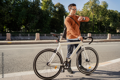 A city dweller pushing a bicycle and using a watch and a phone passes by the park.