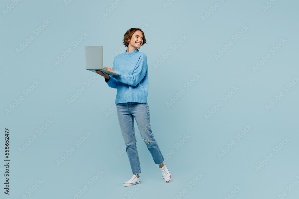 Full body side view young IT woman wear knitted sweater hold use work on laptop pc computer look aside on workspace area isolated on plain pastel light blue cyan background People lifestyle concept