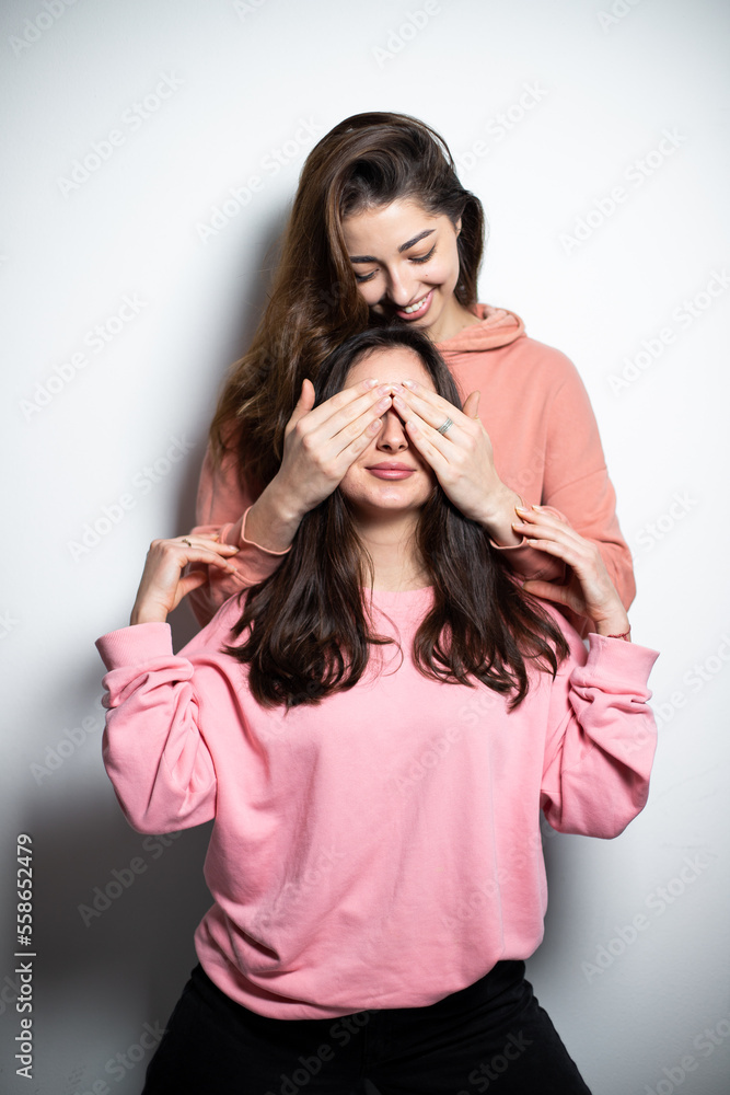 Fototapeta premium Two young attractive brunette girls in pink sweaters smiling, one is covering the eyes of another with hands, making surprise, white background