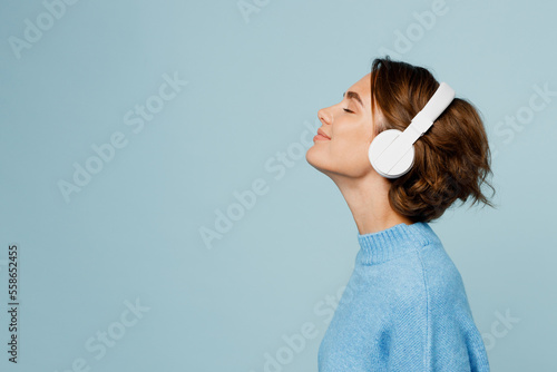 Young happy fun caucasian woman wear knitted sweater headphones listen to music with closed eyes have fun isolated on plain pastel light blue cyan background studio portrait. People lifestyle concept.