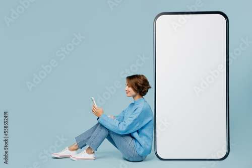 Full body happy sideways young woman in knitted sweater sit near big huge blank screen mobile cell phone with workspace mockup area use smartphone isolated on plain pastel light blue cyan background
