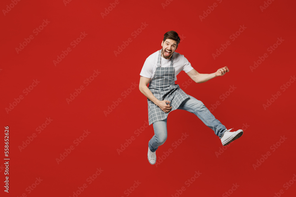 Full body musician fun young male housewife housekeeper chef cook baker man wear grey apron jump high pov playing guitar do hand gesture isolated on plain red background studio. Cooking food concept