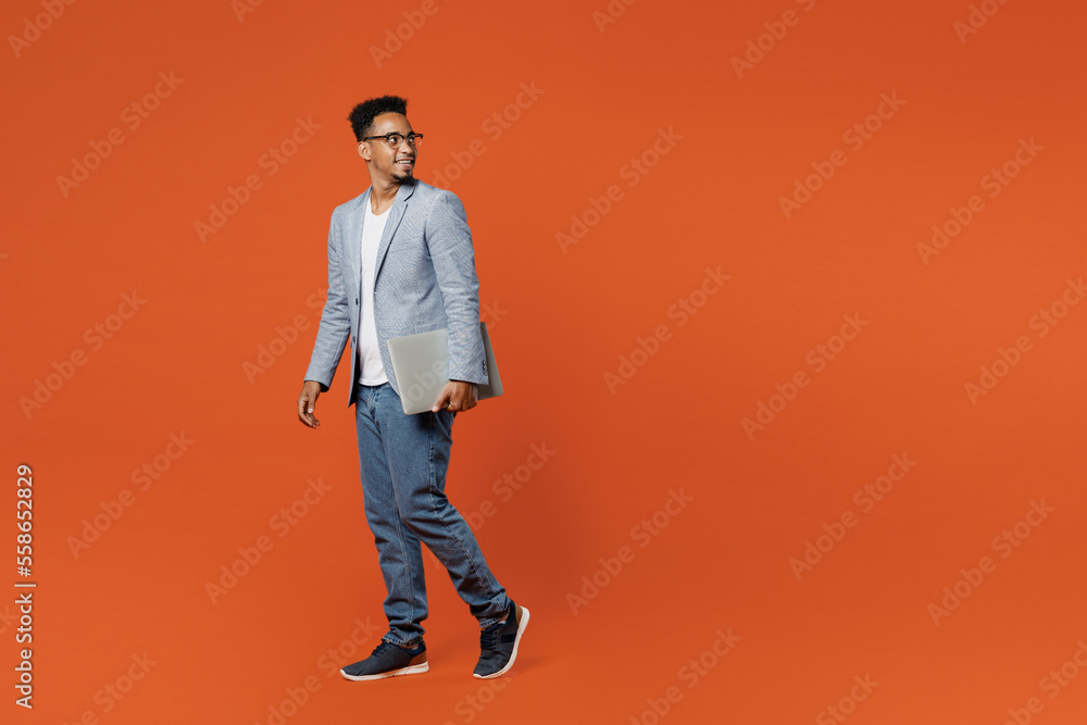 Full body young employee IT business man corporate lawyer wear formal grey suit shirt glasses work in office hold closed laptop pc computer look aside isolated on plain red orange background studio. Full body young employee IT business man corporate lawyer wear formal grey suit shirt glasses work in office hold closed laptop pc computer look aside isolated on plain red orange background studio.