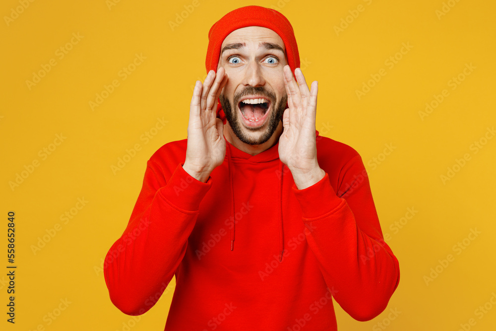 Young promoter caucasian man wear red hoody hat scream sharing hot news about sales discount with hands near mouth isolated on plain yellow color background studio portrait. People lifestyle concept. Young promoter caucasian man wear red hoody hat scream sharing hot news about sales discount with hands near mouth isolated on plain yellow color background studio portrait. People lifestyle concept.