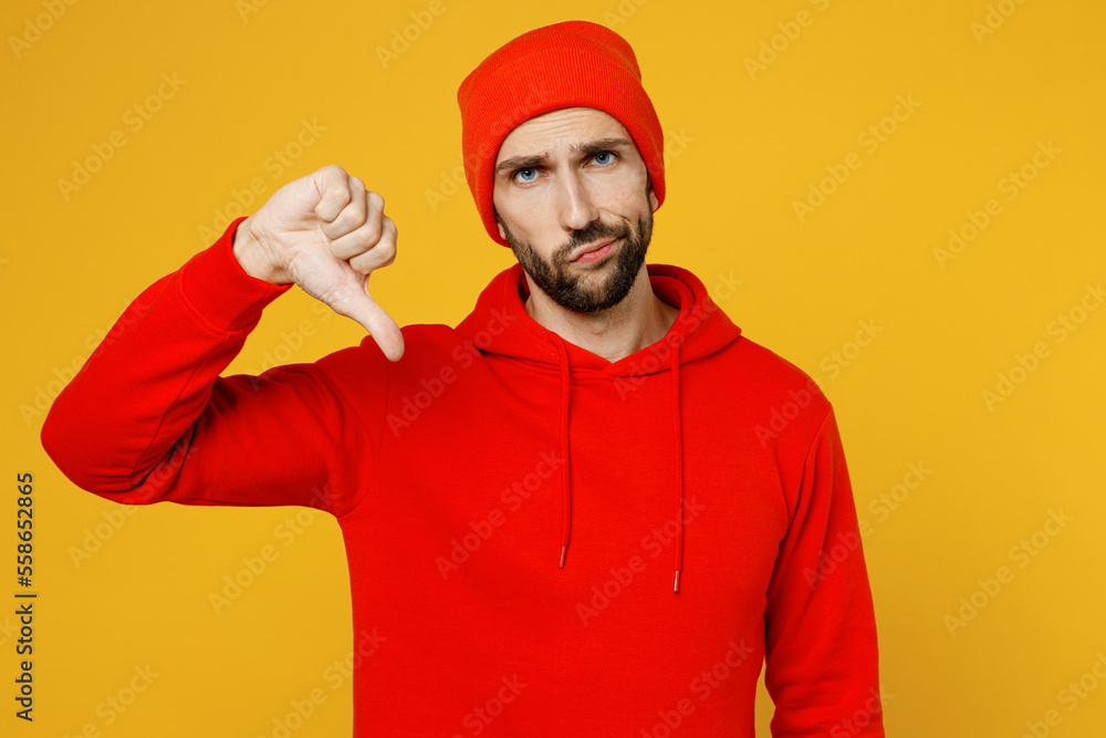 Fototapeta premium Young displeased dissatisfied sad caucasian man wear red hoody hat look camera showing thumb down dislike gesture isolated on plain yellow color background studio portrait. People lifestyle concept.