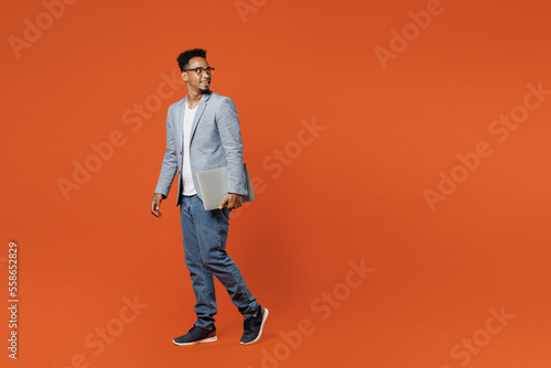 Full body young employee IT business man corporate lawyer wear formal grey suit shirt glasses work in office hold closed laptop pc computer look aside isolated on plain red orange background studio.