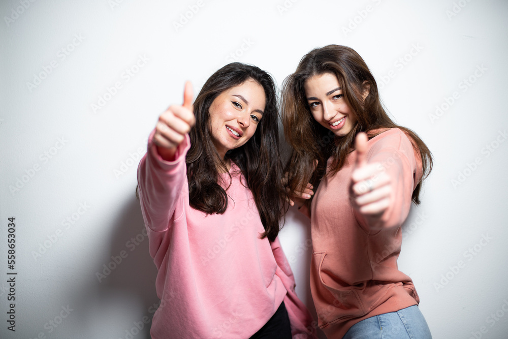Two young attractive brunette girls in pink sweaters, smiling, showing like gesture with fingers, white background Two young attractive brunette girls in pink sweaters, smiling, showing like gesture with fingers, white background