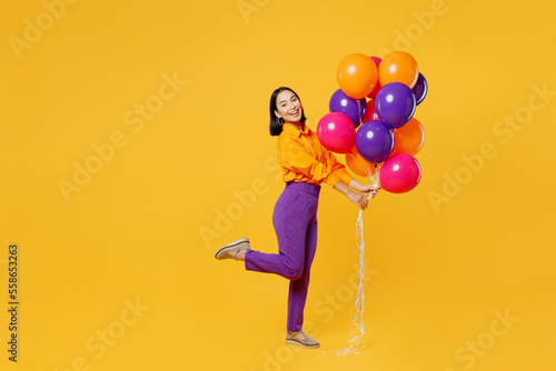 Full body side view smiling happy fun young woman wearing casual clothes hat celebrating hold bunch of balloons raise up leg isolated on plain yellow background. Birthday 8 14 holiday party concept.