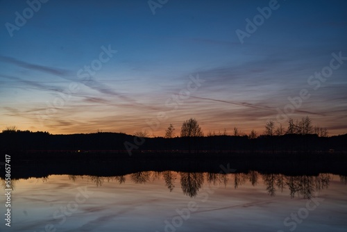 Wonderful blue golden red sky after sunset with trees in foreground at a lake with its reflections 