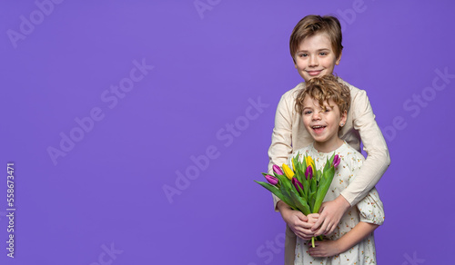 Friendly hugs between cute smile children boy and girl standing over purple background, hold spring tulips, looking at camera. Copy space banner for advertisement.