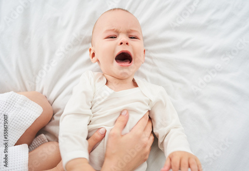 A crying newborn boy in a white bodysuit on a bed.